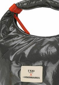 Schwarze glänzende Tasche mit strukturierter Oberfläche, die einen auffälligen orangen Riemen und ein Etikett mit der Aufschrift "UNIO HAMBURG X LES VISIONNAIRES" trägt.