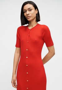 Κόκκινο ριμπ φόρεμα με κοντά μανίκια, διαθέτει στρογγυλή λαιμόκοψη και χρυσά κουμπιά κατά μήκος του μπροστινού μέρους. Ομαλή υφή και στενή εφαρμογή.