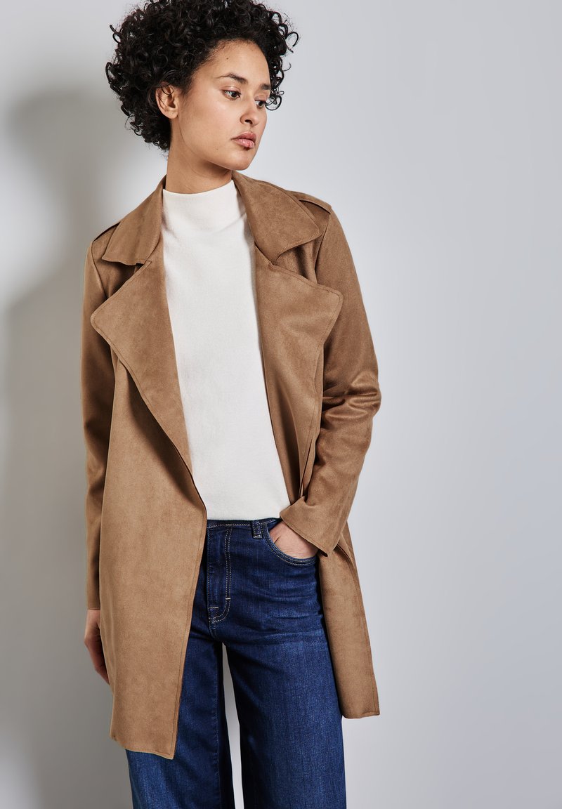 Cappotto trench in suede marrone con ampie rever, indossato sopra un dolcevita bianco. Abbinato a jeans a gamba larga blu. Linee e texture semplici e pulite.