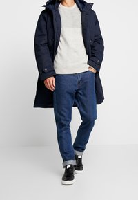 Parka navy con bottoni, maglione grigio a trama, jeans di denim blu e sneaker nere con suole bianche. Abbigliamento casual.