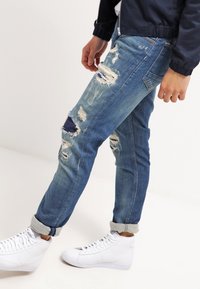 Vaqueros azules desgastados con múltiples roturas, corte ajustado y puños doblados. Combinados con zapatillas blancas de caña alta.