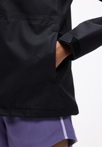 Personne portant une veste noire avec la main dans la poche latérale et un short violet avec une bande violet clair sur fond blanc.