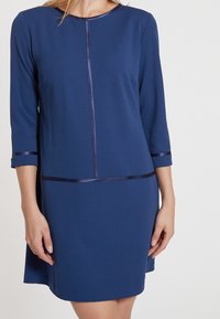 Mörkblå klänning med trekvartsärmar, som har en satinbård längs halsringningen och ärmarna, slät tyg och en rak silhuett.