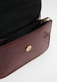 Burgundská umělá kůže taška s krokodýlím vzorem, se zlatým zapínáním na knoflík a černým podšívkou s kapsou na zip.