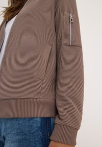 Bruine geribde jas met een gestructureerd oppervlak, voorzien van een ritszak op de mouw en een aansluitende geribde zoom. Gecombineerd met blauwe jeans.