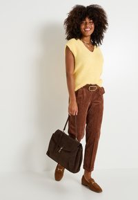 Top sans manches en tricot jaune duveteux, pantalon en velours côtelé marron taille haute, mocassins en daim marron et un sac à main en cuir marron avec des accents dorés.