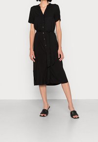 Schwarzes, kurzärmeliges Kleid mit Knopfverschluss, tailliertem Gürtel, Midi-Länge und seitlichen Schlitzen. Getragen mit schwarzen, gewebten Rutschschlappen auf einem hellgrauen Boden.