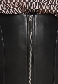 Close-up van een zilveren rits op een zwart leren kledingstuk onder een patroon van zwart-wit stof.