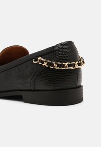 Mocassin en cuir noir texturé avec un petit talon et un détail décoratif en chaîne dorée à l'arrière.