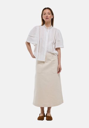 Femme portant un chemisier blanc à manches courtes avec des détails en dentelle, une longue jupe beige avec des poches devant, et des chaussures plates marron, debout sur un fond blanc.