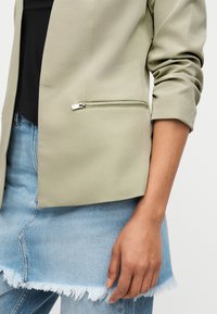 Personne portant un blazer vert clair avec une poche zippée, un haut noir, une jupe en jean bleu à ourlet effiloché et un jean.