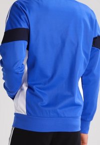 Blå idrottsjacka med en svart axelrand, vita sidopaneler och en slät textur. Designad med en rundad krage och åtsittande ärmmuddar.