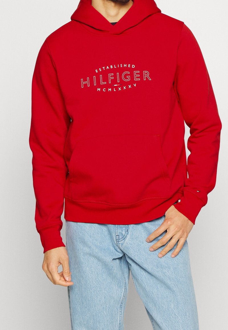 Felpa rossa con cappuccio realizzata in tessuto morbido, con tasca frontale e testo con logo bianco, indossata con jeans azzurri chiari.