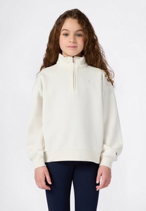 Unge pige med langt krøllet hår iført en hvid lynlås sweatshirt og mørkeblå bukser, der står foran en ensfarvet hvid baggrund.