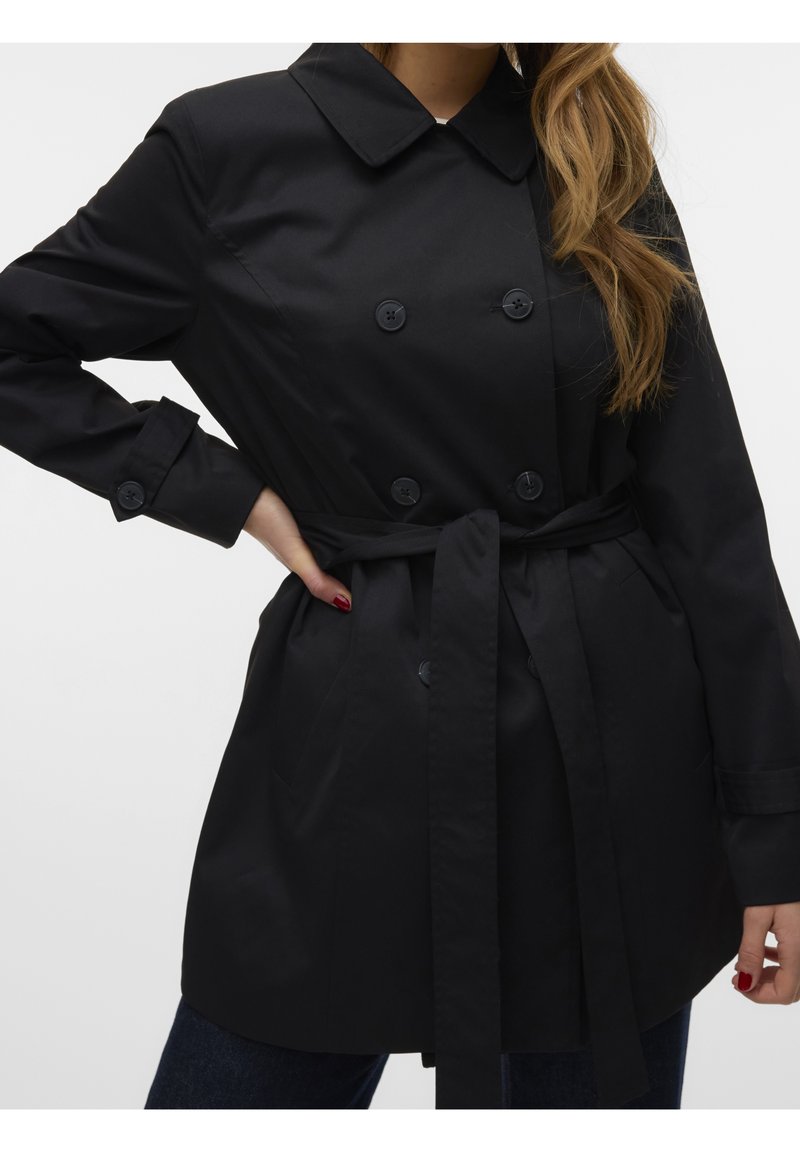 Femme portant un trench-coat noir croisé avec ceinture à nouer, mains sur les hanches, et longs cheveux ondulés partiellement visibles.