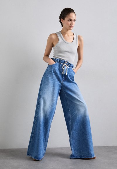 Φαρδείς μπλε τζιν παντελόνες με φθαρμένη εμφάνιση, διαθέτοντας ελαστική μέση με κορδόνι και πλαϊνές τσέπες. Συνοδεύονται από ένα γκρι αμάνικο τοπ.