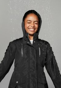 Giacca nera impermeabile con zip frontale, due tasche sul petto e cappuccio. Dettagli testurizzati sulle spalle con gocce d'acqua visibili.