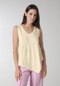 Top galben deschis, fără mâneci, cu decolteu în V, la o linie asimetrică a tivului și un material ușor texturat. Asortat cu pantaloni roz.