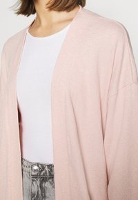 Kimono de color rosa claro, abierto por delante, con tejido texturizado, usado sobre una blusa ajustada blanca, combinado con pantalones metálicos gris de cintura alta.