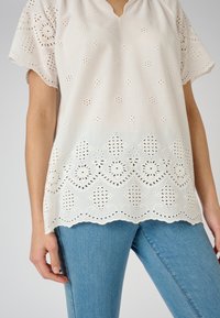 Camicia bianca con ricamo a occhio e orlo scallop, dotata di maniche corte e vestibilità comoda. Abbinata a jeans azzurri.
