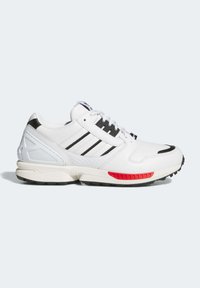 Zapatilla deportiva blanca con rayas negras, parte superior de cuero texturizado, cuello acolchado, acento rojo en la suela y suela de goma con patrón de tracción.