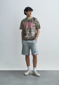 Γραφικό μπλουζάκι με καφέ και πράσινο μοτίβο, με έντονο ροζ κείμενο· συνδυασμένο με γκρι σορτς και άσπρα αθλητικά παπούτσια.