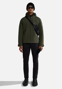 Giacca impermeabile verde oliva con cappuccio, borsa a tracolla nera, pantaloni neri e stivali neri. Il modello indossa occhiali da sole e un berretto.