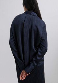 Camisa de rayas finas azul marino con una textura suave, mangas largas y puños abotonados, vista desde atrás con las manos entrelazadas detrás.
