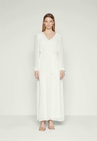 Γυναίκα που στέκεται φορώντας ένα μακρύ λευκό φόρεμα με V λαιμόκοψη, μακριά μανίκια, ζώνη στη μέση, και μπεζ σανδάλια με λουριά σε απλό ανοιχτό φόντο.