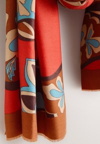 Tissu coloré suspendu, présentant des motifs floraux audacieux en bleu, marron, rouge et beige, avec une texture douce et des bords frangés.