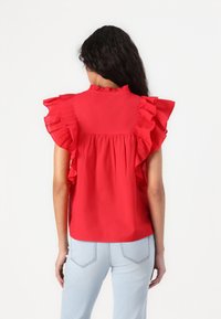 Blouse rouge avec manches à volants et col montant. Le tissu est léger, lisse et présente un design froncé au dos. Associée à un jean bleu clair.