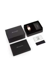 Rektangulært guldfarvet armbåndsur med brun læderrem i sort fløjlæske, med to sorte yderkasser, instruktionskort og renseklud.