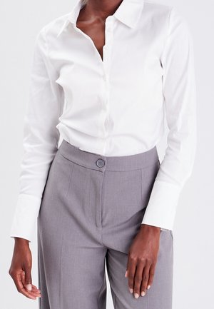 Personne portant une chemise blanche boutonnée rentrée dans un pantalon gris taille haute avec une fermeture à un seul bouton.