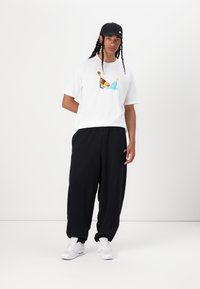 Model i en hvid bomulds t-shirt med et farverigt grafisk motiv, parret med løse sorte bukser og hvide sneakers. En sort kasket fuldender outfittet.