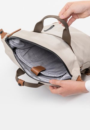 Handen die een beige stoffen rugzak openen met daarin een grijs geruit gewatteerd laptopvak en een bruine leren riem.