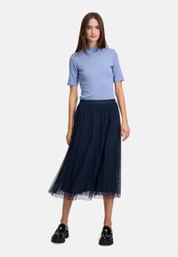 T-shirt fitted blu chiaro a maniche corte abbinato a una gonna midi in tulle blu navy scuro. La modella indossa scarpe con plateau nere; design minimalista.
