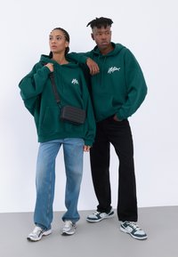 Grønne hættetrøjer med hvidt logo, parret med lyse denim og sorte bukser. Den ene bærer en sort crossbody taske; begge har atletiske sko på.