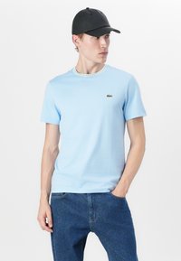 T-shirt bleu clair à manches courtes avec un col côtes, arborant des accents verts et jaunes, associé à un jean en denim bleu. Logo Lacoste sur la poitrine.