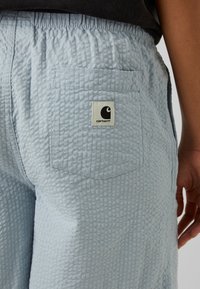 Ljusblå, texturerade byxor med en rynkad midja och bakfickor. Har en synlig Carhartt-logotyp på fickan.