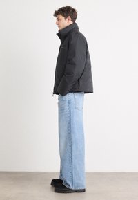 Zwarte pufferjas met hoge kraag, gedragen over lichtblauwe wijde spijkerbroek. Zwarte chunky schoenen maken de outfit compleet. Effen achtergrond.