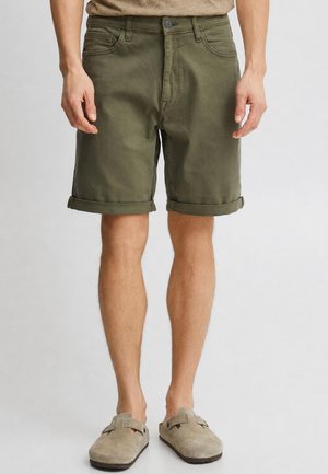 Homme portant un short kaki avec revers et des chaussures beige sans lacets, debout devant un fond clair uni.