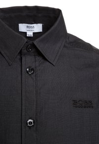 Camicia nera con bottoni, caratterizzata da un colletto corto, una texture discreta e un logo ricamato sul lato sinistro del petto. Ha bottoni neri e una vestibilità regolare.