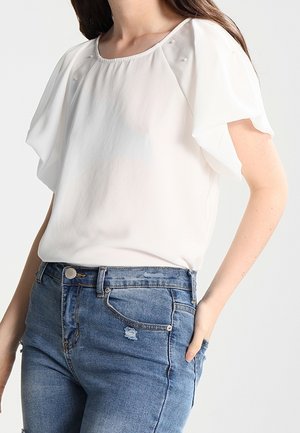 Valkoinen lyhythihainen paita, jossa röyhelöhihat, yhdistetty korkeavyötäröisiin sinisiin farkkuihin, käsi rentosti taskussa.