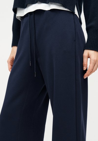 Κοντινό πλάνο ενός ατόμου που φοράει σκούρο μπλε παντελόνι με φαρδύ πόδι και ζωνάκι στη μέση, και μαύρο μακρυμάνικο πάνω από λευκό πουκάμισο.