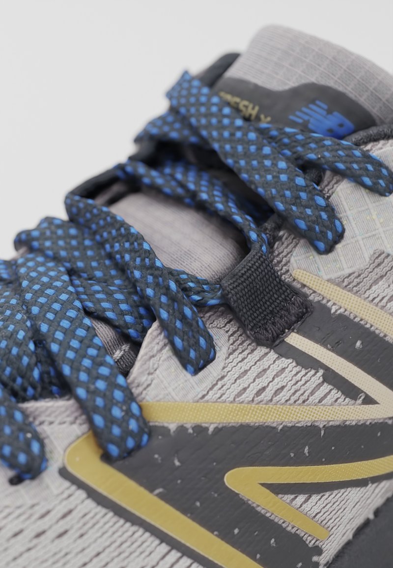 Zapatilla deportiva gris con una parte superior texturizada, acentos en negro y dorado, y cordones de malla azul. Cuenta con una lengüeta acolchada y una zona de puntera reforzada.