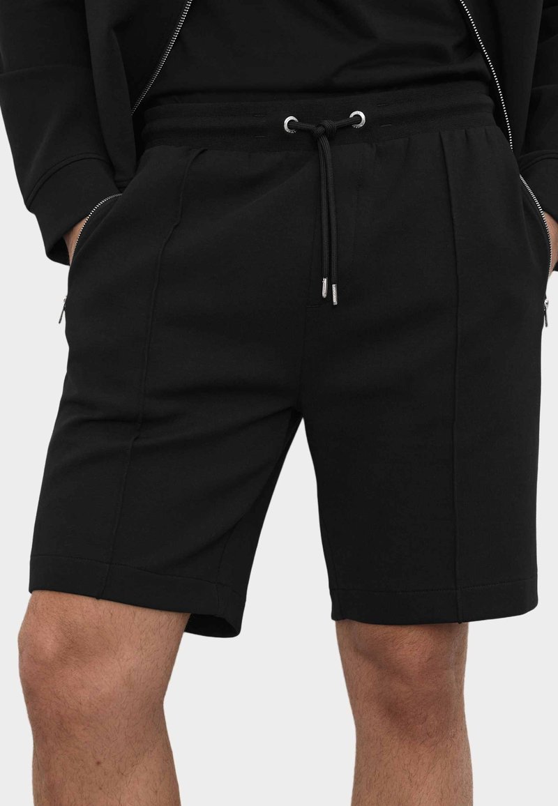 Black mid-length shorts with drawstring waist, side zip pockets, worn with hands in pockets visible on sides.