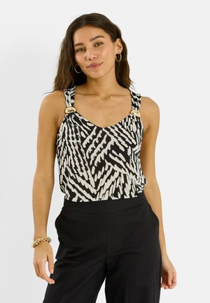 Femme portant un haut sans manches à motifs noir et blanc avec des boutons dorés et un pantalon noir, accessoirisée de boucles d’oreilles créoles dorées et d’un bracelet.