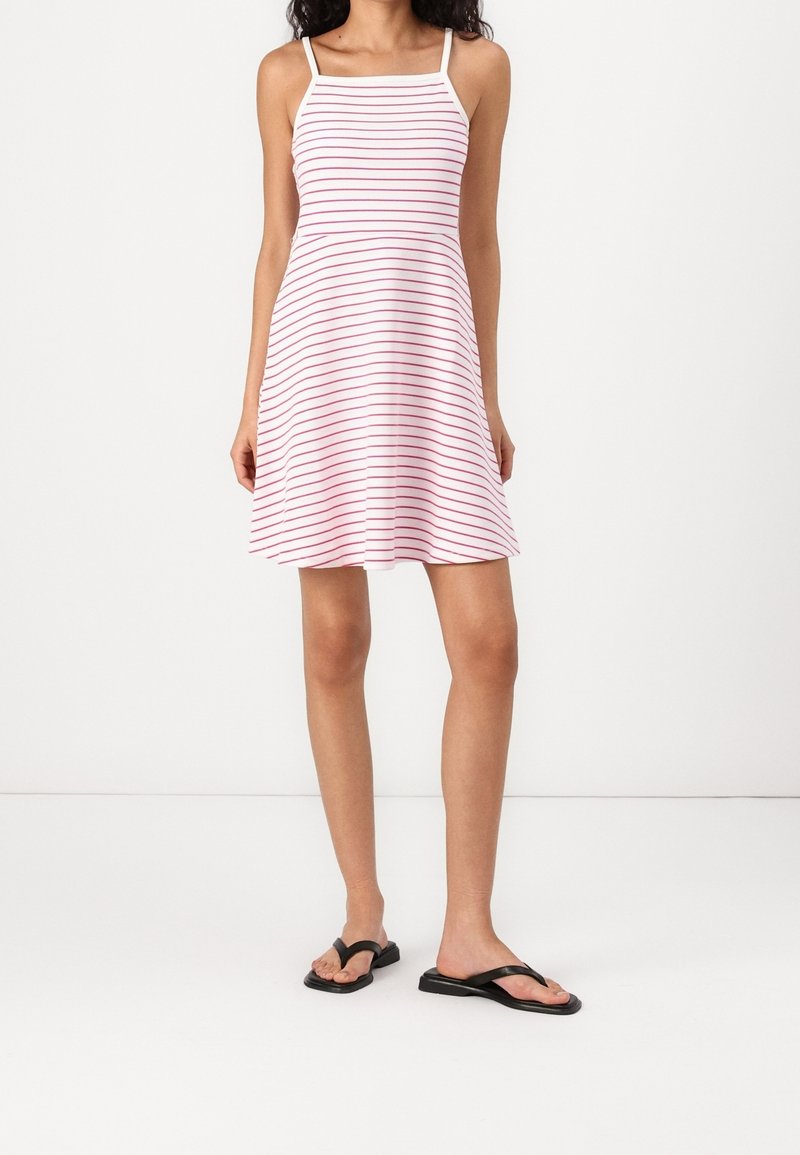 Femme portant une robe blanche sans manches avec de fines rayures rouges horizontales et des sandales plates noires, debout devant un fond uni.