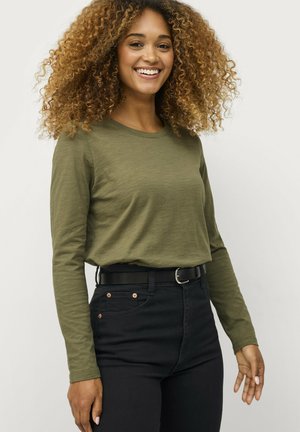 Vrouw met krullend haar die een olijfgroene lange mouwen shirt en hoge taille zwarte broek draagt, lachend tegen een effen achtergrond.