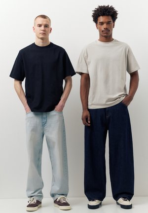 Két férfi áll egymás mellett fehér háttér előtt, sima, túlméretezett pólót és bő szárú farmert viselve, kezük a zsebekben.